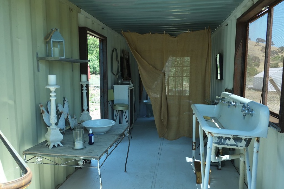 Een rustieke buitenbadkamer in een container-achtige structuur met grote ramen die uitzicht bieden op de natuur, ingericht met vintage sanitair en natuurlijke materialen.
